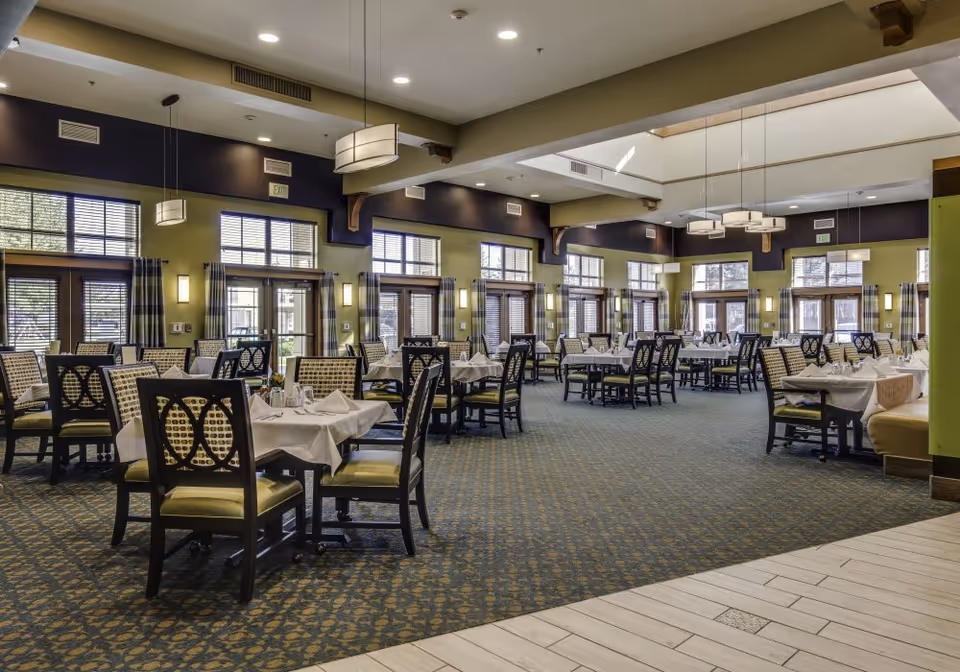 Spacious dining room with multiple tables set with white linens and chairs, large windows and pendant lighting.