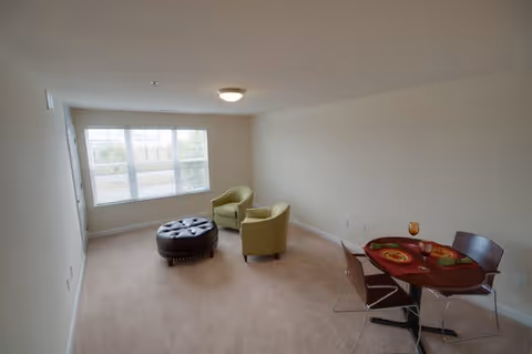 A small living area with beige carpet and walls, featuring two green armchairs and a round black ottoman near a large window. A small round dining table with two place settings and two chairs is positioned against the right wall.