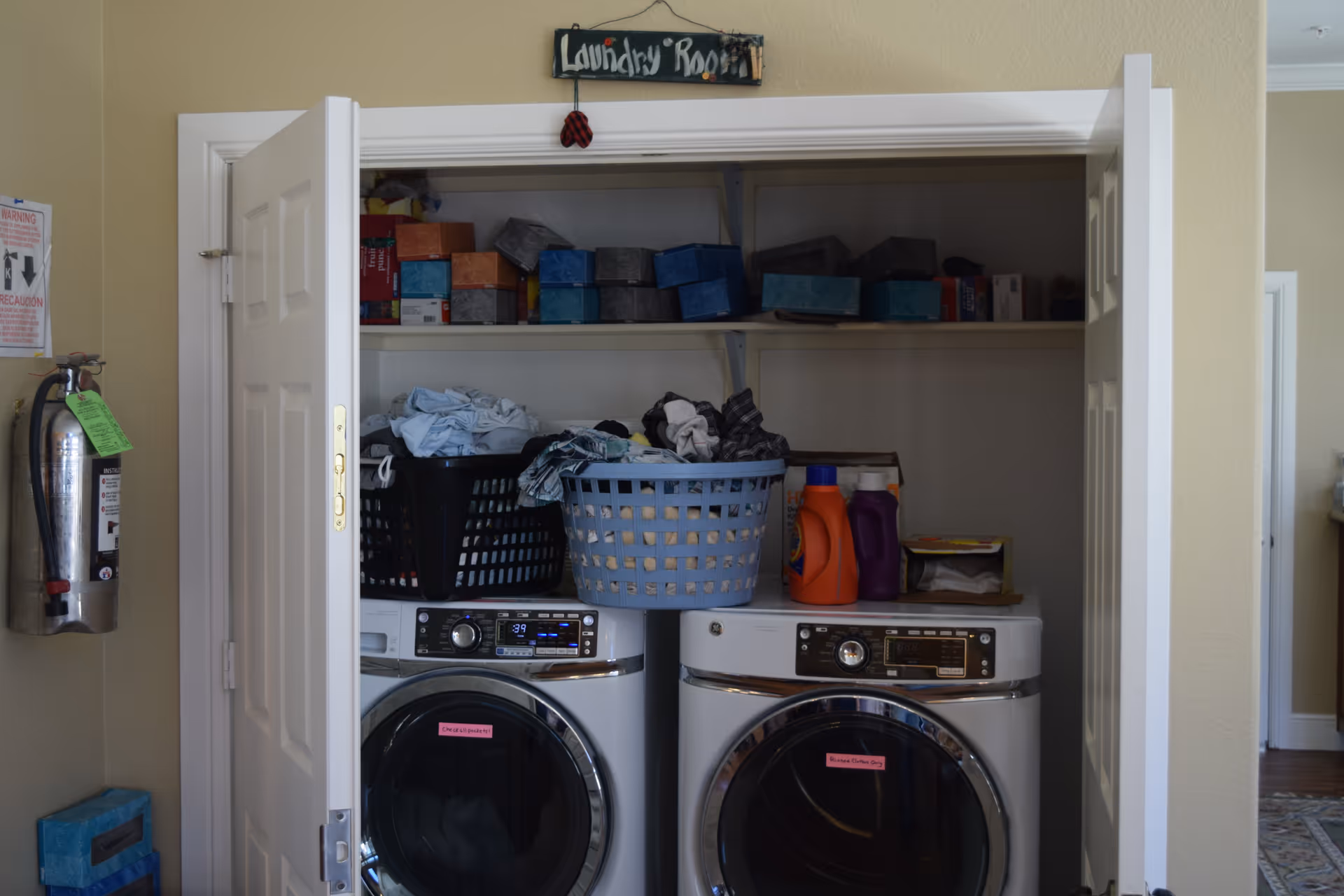 A laundry room with two front-loading washing machines side by side. Above the machines are two laundry baskets filled with clothes and shelves holding various boxes and laundry supplies. The room is labeled with a small sign above the door that reads 'Laundry Room'. A fire extinguisher is mounted on the wall to the left of the door.