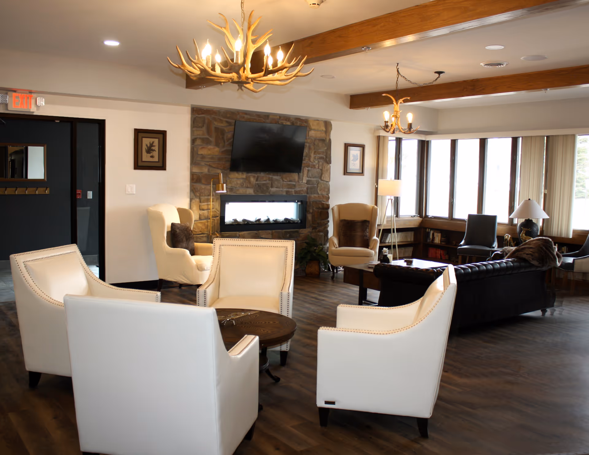 A spacious communal living room with white armchairs around a coffee table, a stone fireplace with a mounted TV, and large windows letting in daylight.