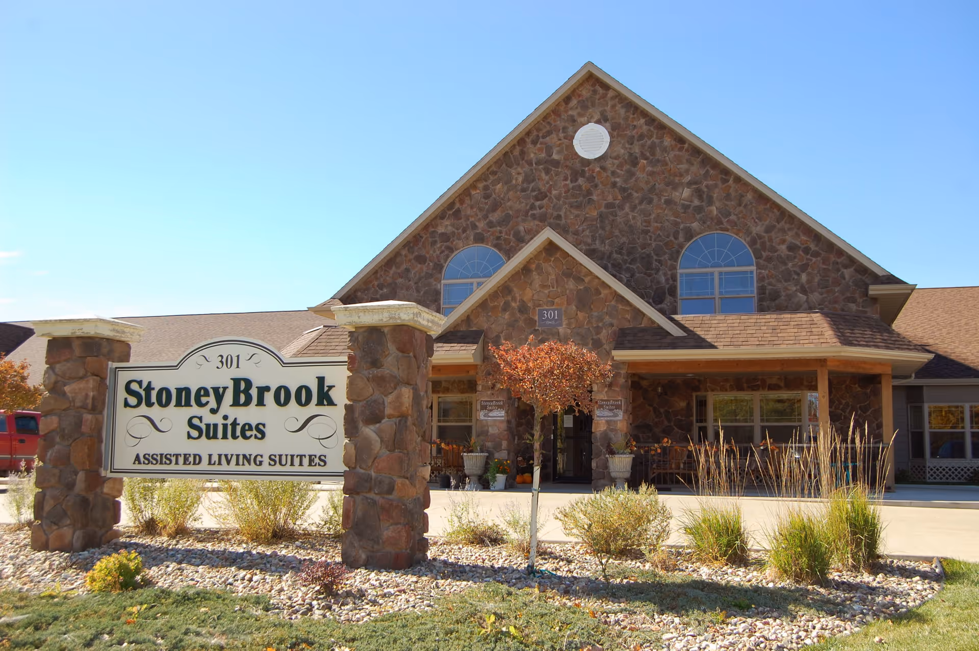 Exterior view of StoneyBrook Suites Assisted Living building with a stone facade, a peaked roof, and a sign in front displaying the facility name and address number 301. The area around the sign has landscaping with small bushes and rocks.