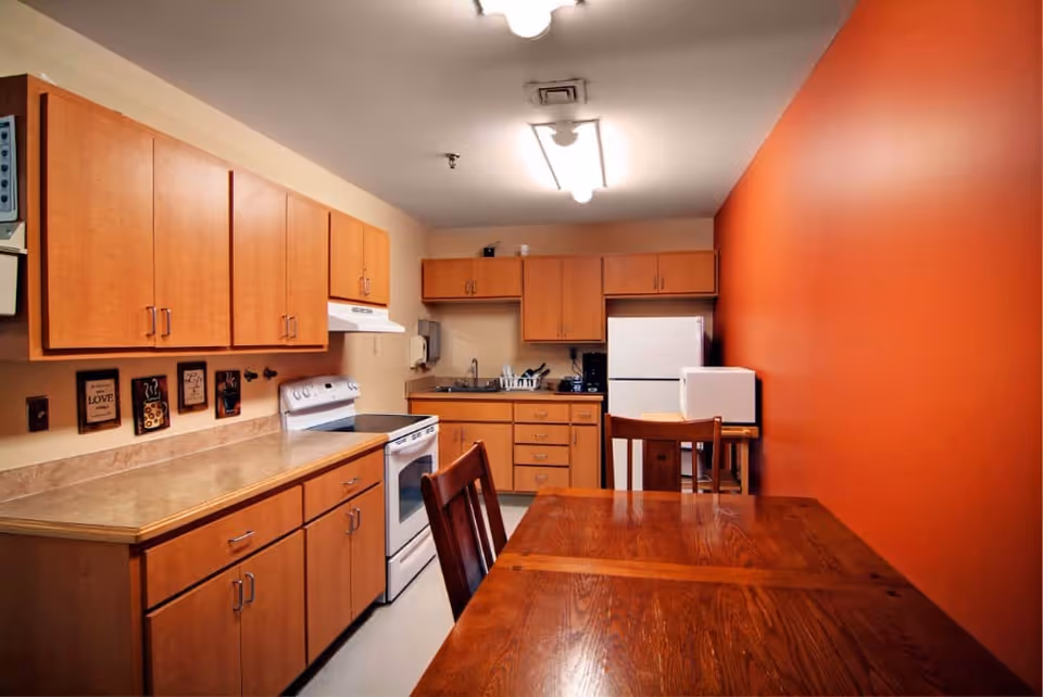 Small communal kitchen with wooden cabinets, white appliances and a wooden dining table against an orange accent wall.