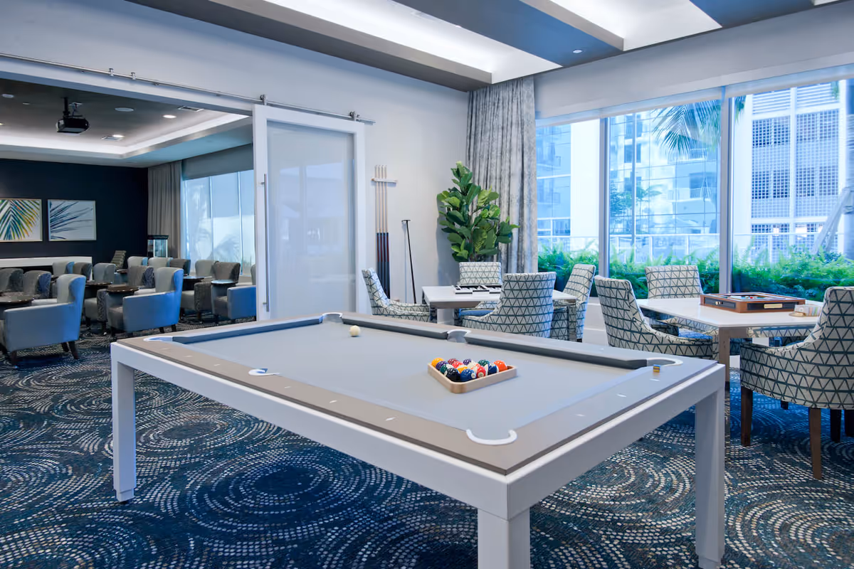 Bright communal game room with a pool table, multiple seating areas, and large windows overlooking greenery.