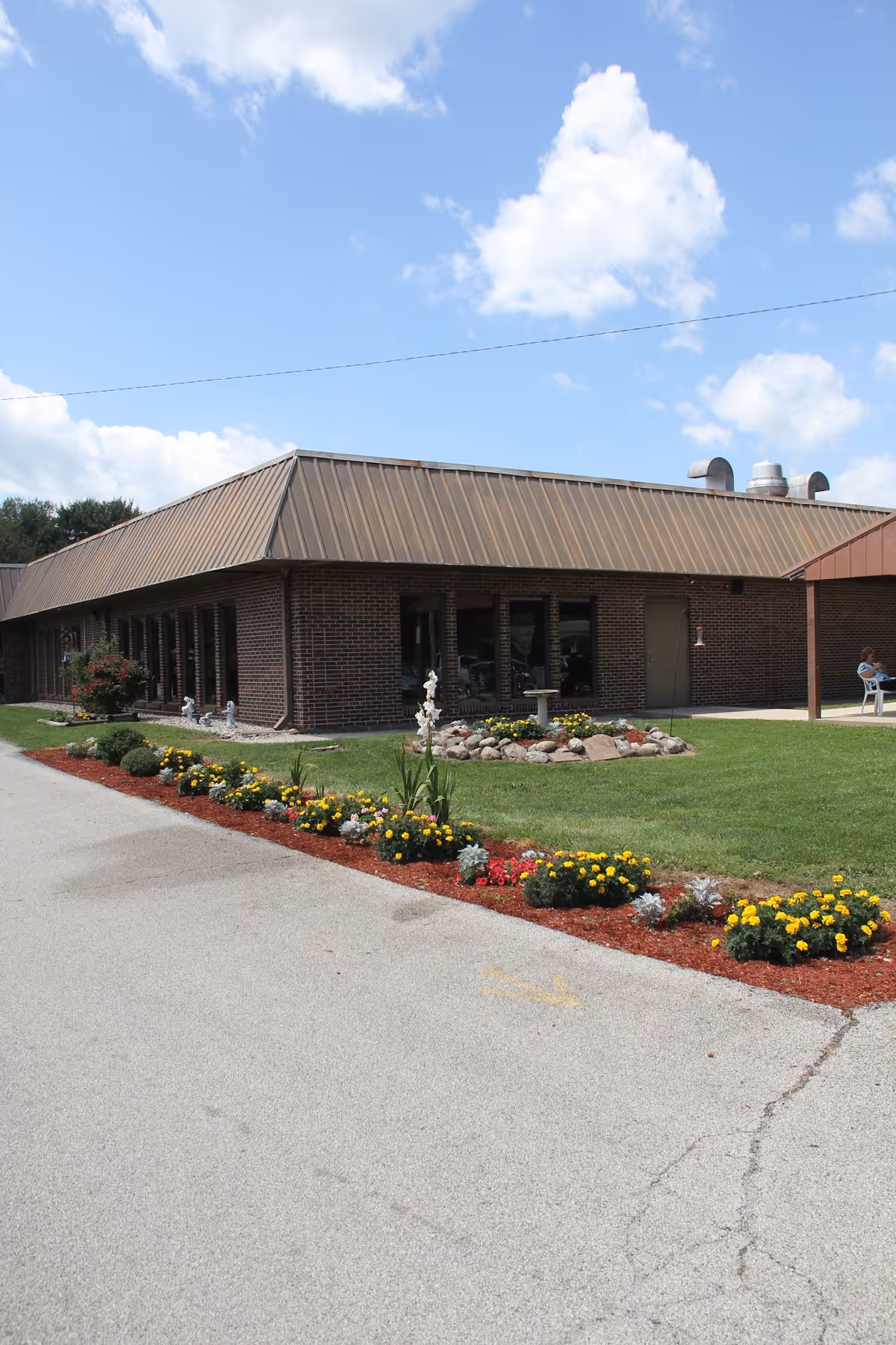 Exterior view of a single-story brick building with a brown metal roof under a partly cloudy sky. The building is surrounded by a well-maintained lawn and a flower bed with yellow and red flowers along a paved driveway. A person is sitting on a bench near the building entrance.