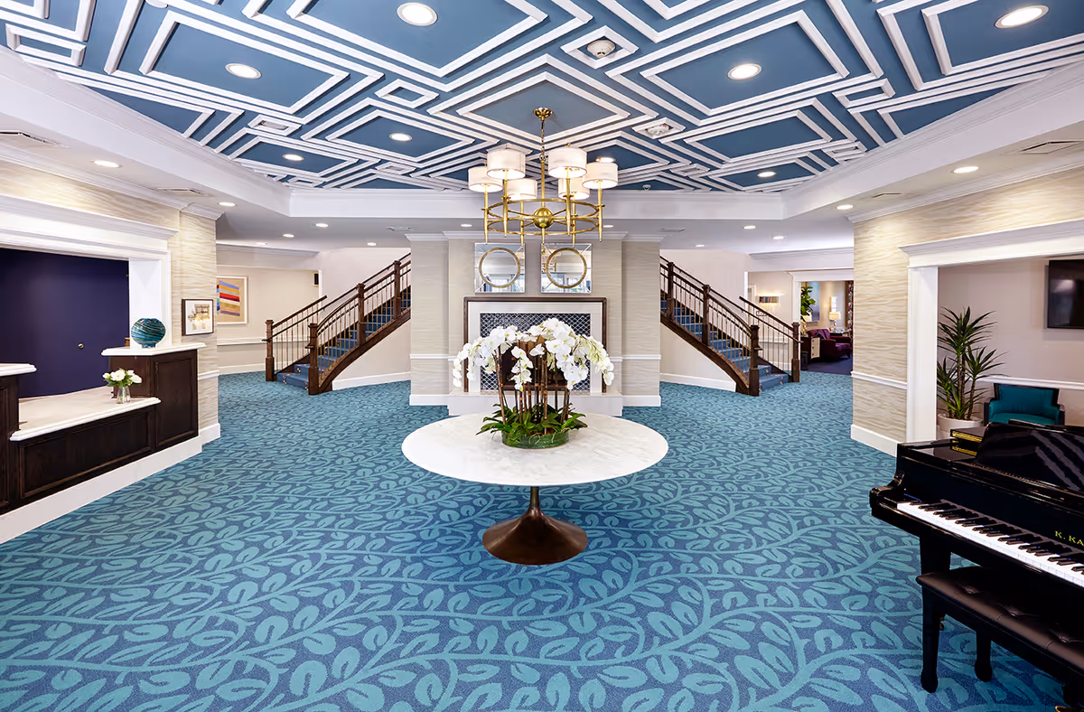 Spacious and elegant lobby area with a patterned blue carpet and a decorative ceiling. A round table with white orchids is centered in the room. Two staircases with wooden railings lead upward on either side of a central fireplace with two round mirrors above it. To the right, there is a black grand piano and some seating with plants. The reception desk is on the left side with a decorative vase and flowers.