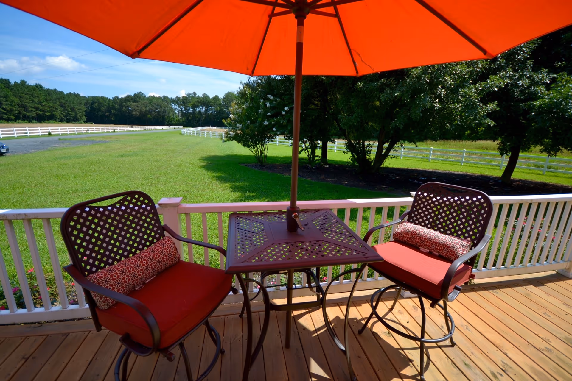 Outdoor patio area with two metal chairs with red cushions and patterned pillows, a square metal table, and a large red umbrella providing shade. The patio overlooks a green lawn with white fencing and trees in the background under a blue sky.