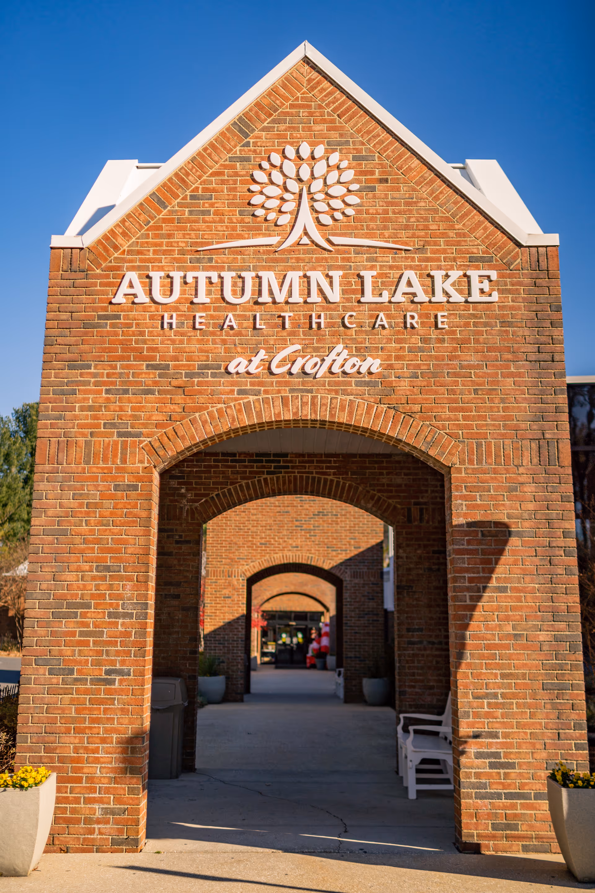 Brick archway entrance with the sign 'Autumn Lake Healthcare at Crofton' displayed prominently above. The archway leads to a walkway with additional brick arches and some potted plants and benches along the sides. The sky is clear and blue.