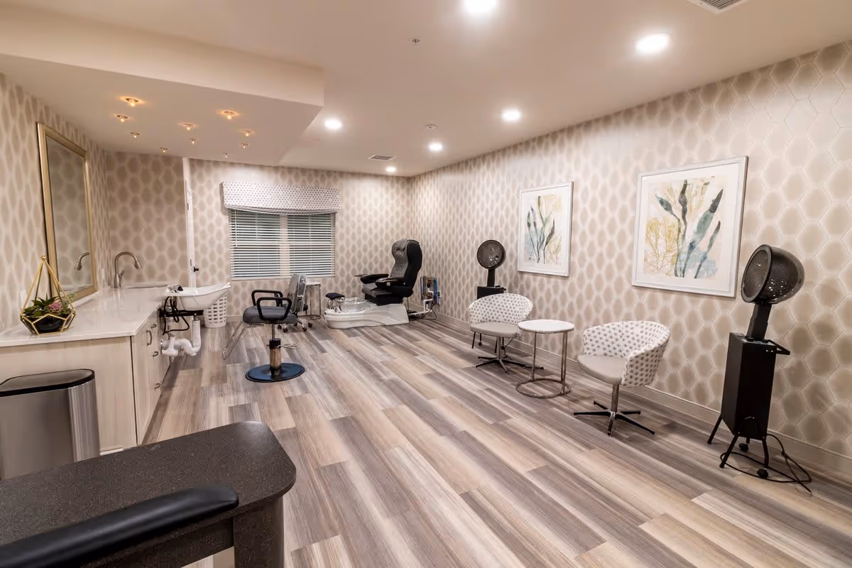 A spacious salon room with light wood flooring and patterned wallpaper. The room features a large mirror above a countertop with a sink, a black salon chair, a pedicure chair with a foot bath, two white patterned chairs with a small round table between them, and two black hair dryers on stands. Two framed botanical artworks hang on the wall.
