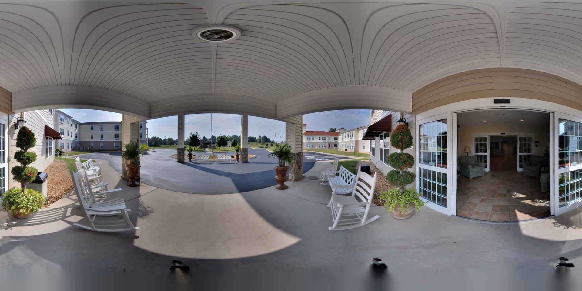Covered main entrance/portico with white rocking chairs, potted plants, a circular driveway and open double doors into the lobby.