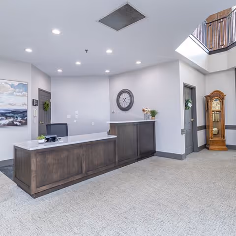 Reception area with a wooden front desk, a black office chair, a wall clock, a painting of a seascape, a grandfather clock, and two doors. The space is well-lit with recessed ceiling lights and has light-colored walls and carpeted flooring.