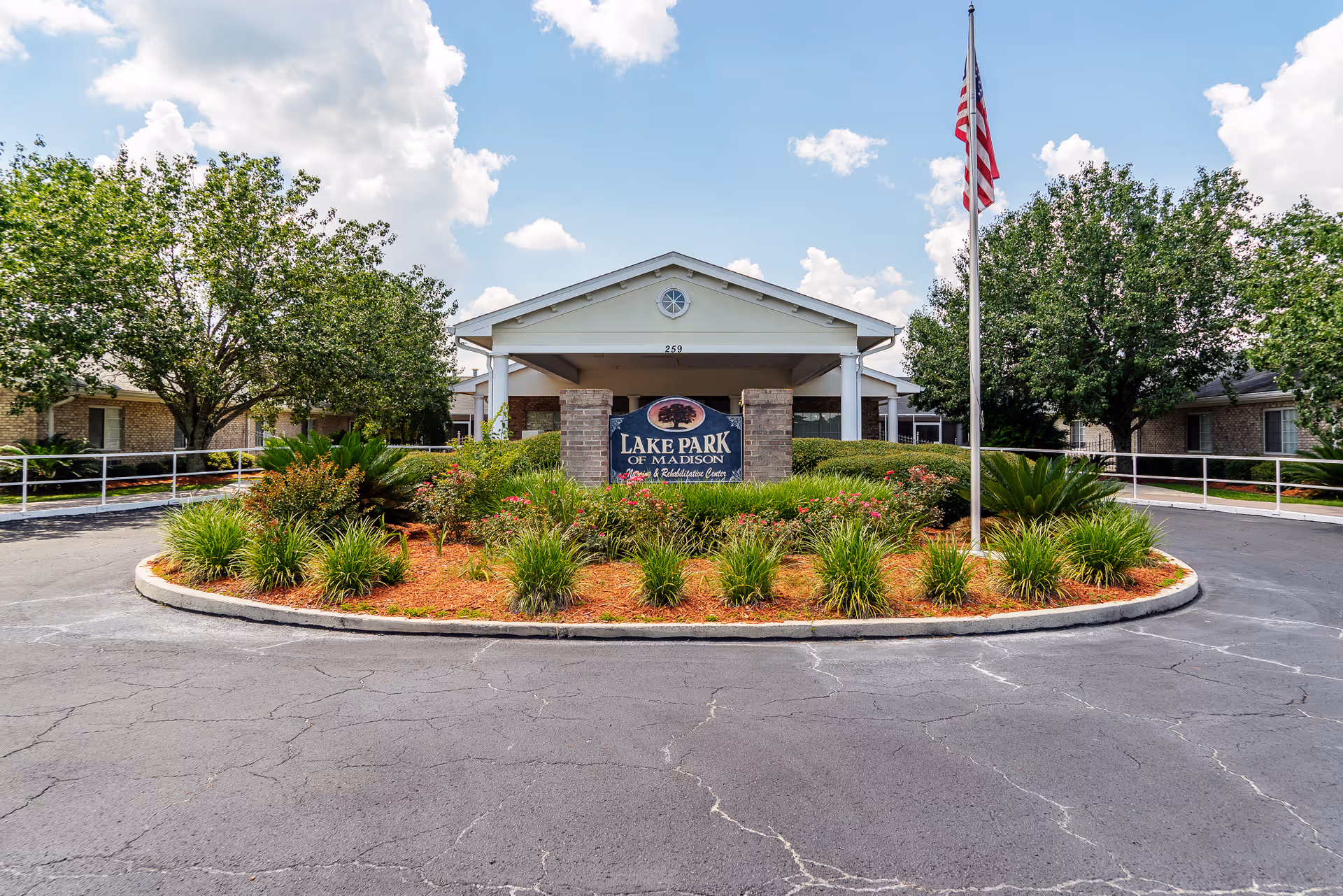 Front exterior view of Lake Park of Madison Nursing and Rehabilitation Center with a circular driveway, landscaped garden with green shrubs and flowers, an American flag on a flagpole, and a partly cloudy sky.