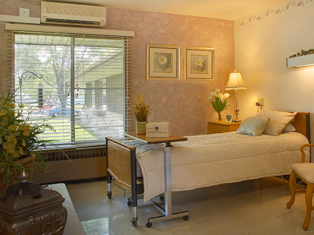 A neatly made single bed in a well-lit room with a window showing an outdoor view. The room has a bedside table with a lamp and flowers, two framed floral pictures on the wall, a chair, and a small table on wheels positioned over the bed.