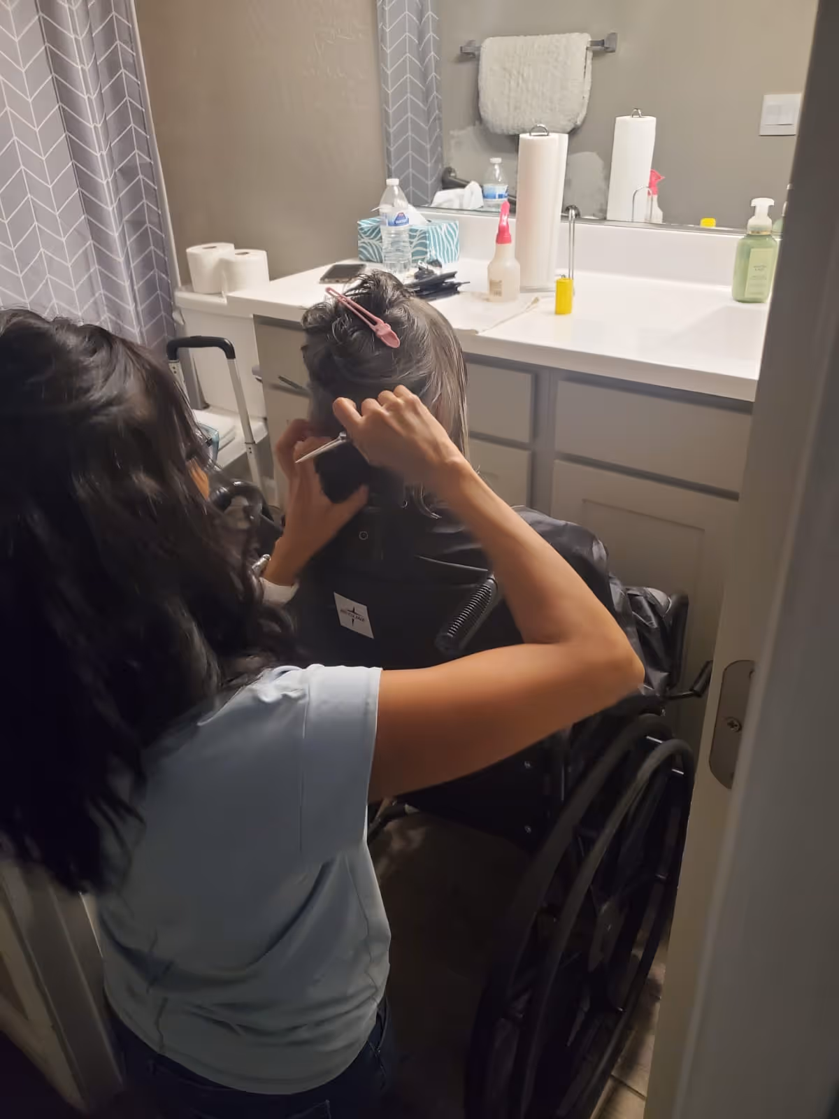 A caregiver trims a person's hair while the person sits in a wheelchair facing the bathroom sink and mirror.