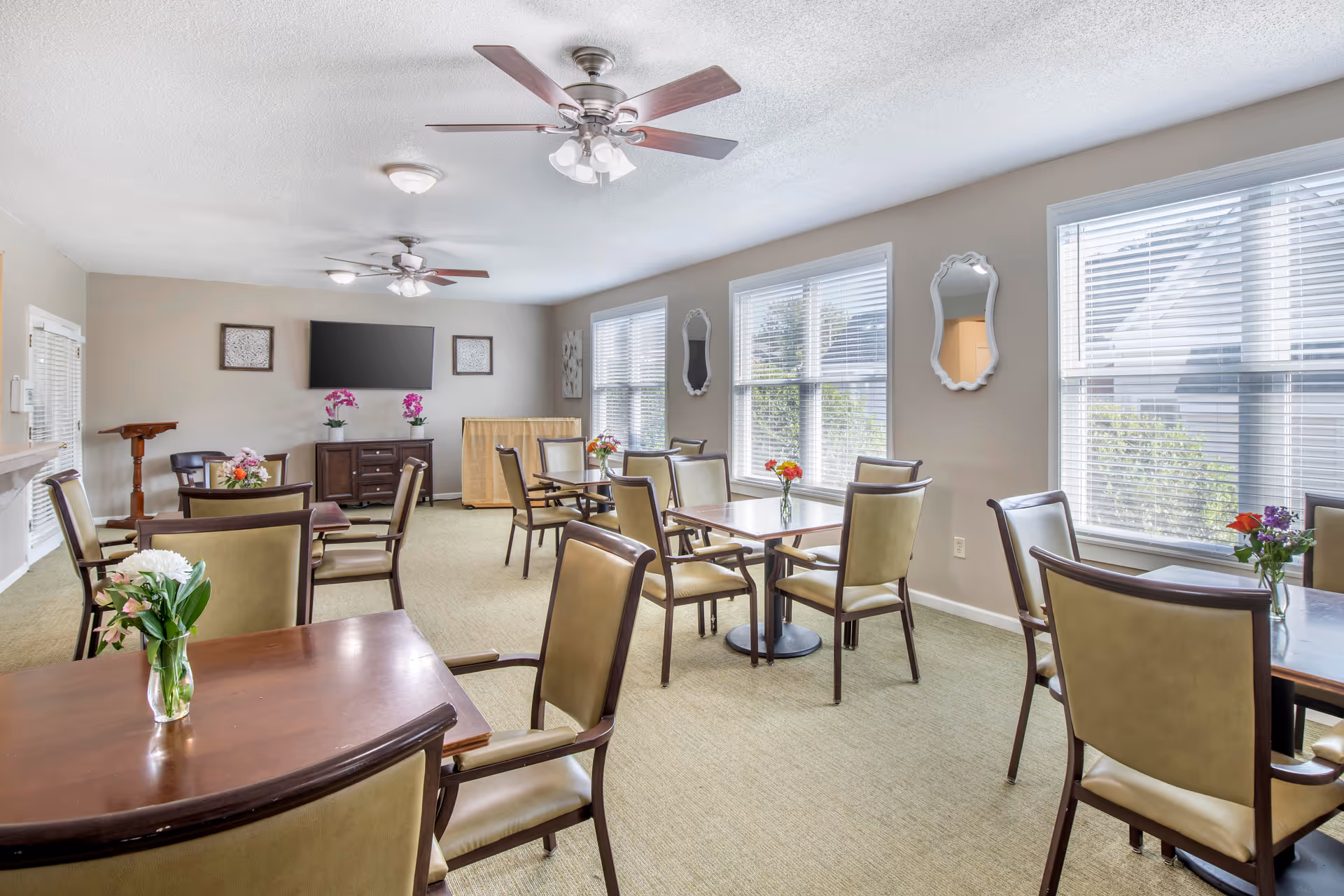 A bright dining room with several wooden tables and beige cushioned chairs arranged neatly. Each table has a small vase with colorful flowers. The room has large windows with white blinds letting in natural light, two ceiling fans with lights, a wall-mounted flat-screen TV, and decorative mirrors on the walls.