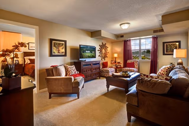 A cozy living room with beige walls and carpet, featuring a brown sofa, two patterned armchairs, a wooden coffee table, and a TV on a wooden cabinet. There are framed pictures on the walls, a window with red curtains letting in natural light, and a glimpse into a bedroom with a bed and nightstand.