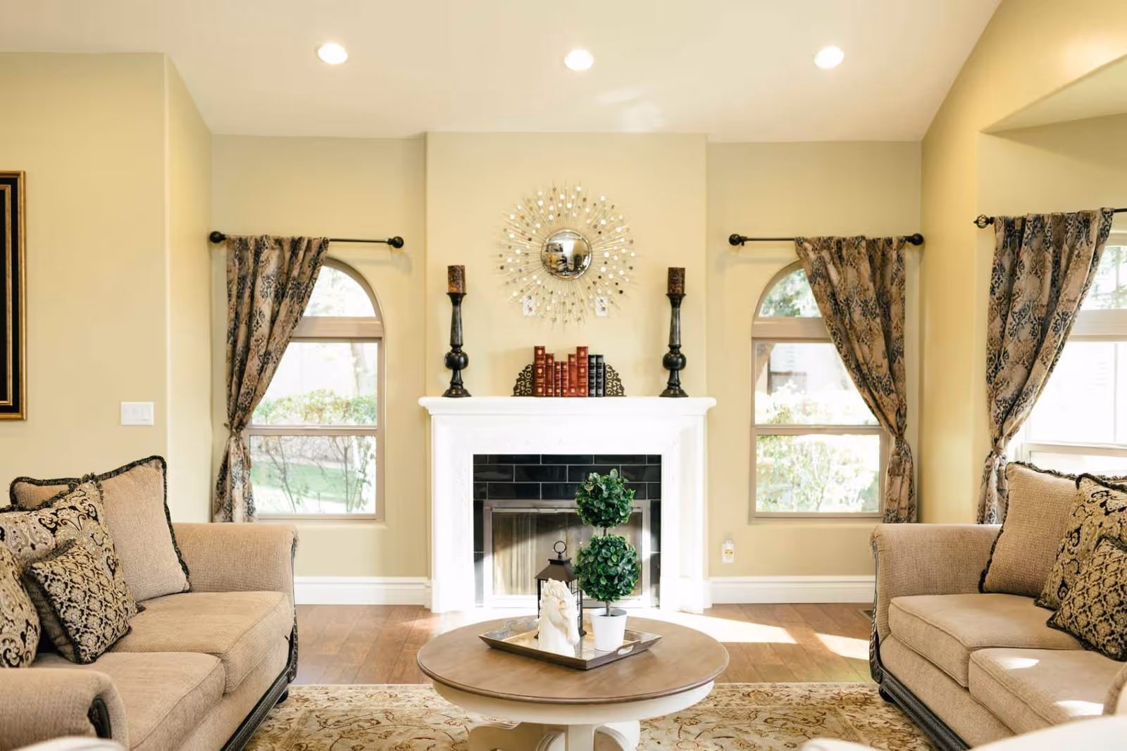 A cozy living room with two beige sofas facing each other, each adorned with patterned cushions. Between them is a round wooden coffee table with decorative plants and a tray. The room features a white fireplace with black tiles, flanked by two tall candle holders and a set of books on the mantel. Above the fireplace is a decorative sunburst mirror. The walls are light beige, and there are three windows with arched tops and patterned curtains allowing natural light to fill the space.