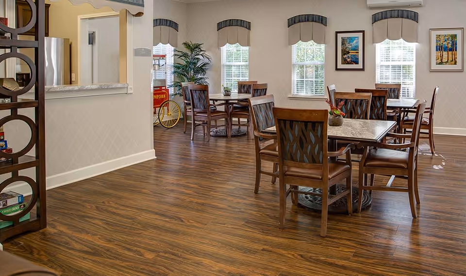 A bright dining area in a senior living facility with wooden floors, several tables and chairs, large windows with valances, framed artwork on the walls, and a popcorn machine in the corner near a potted plant.