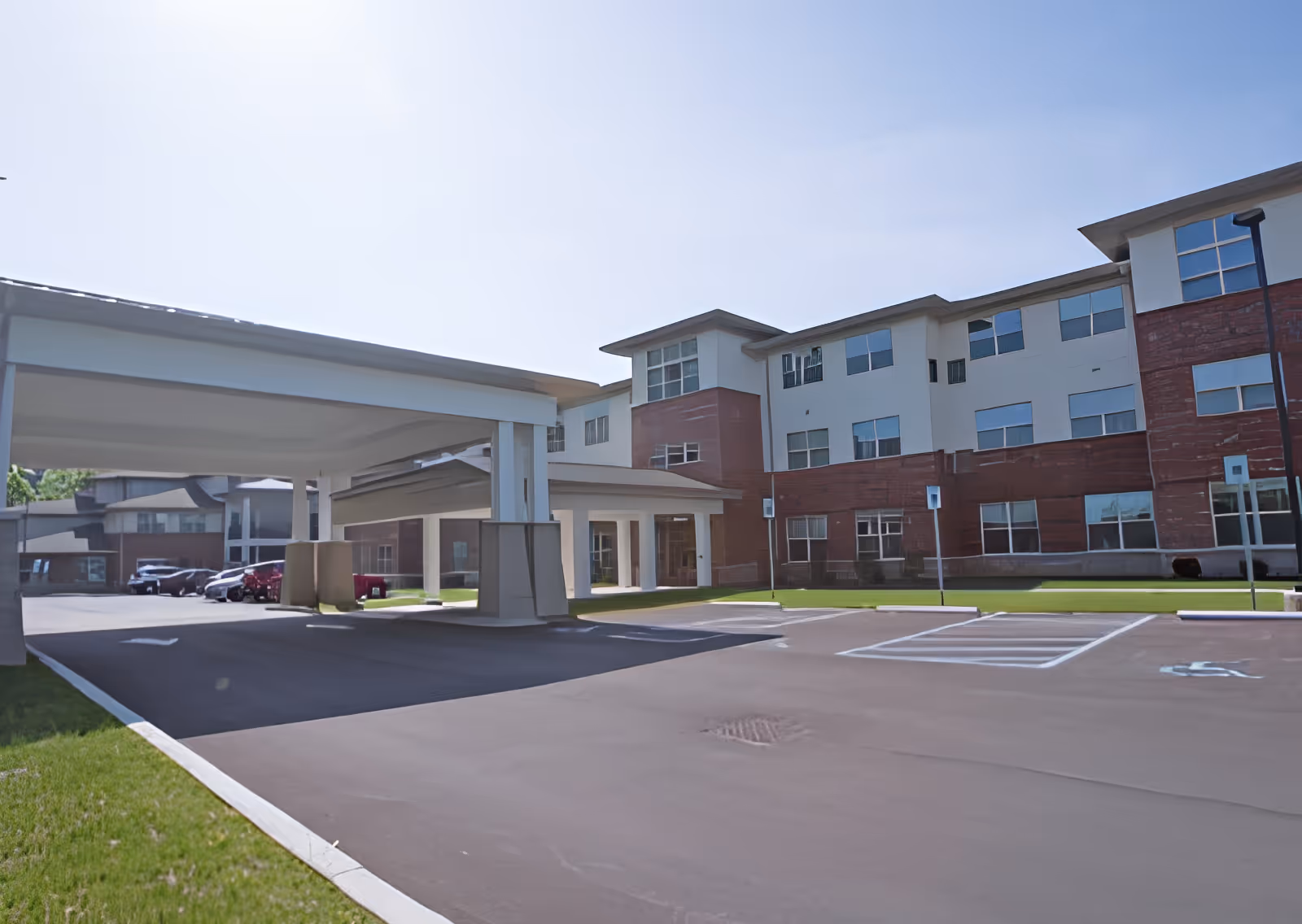 Exterior view of a multi-story senior living facility building with a covered entrance driveway and several parked cars. The building has a combination of brick and light-colored siding with multiple windows.