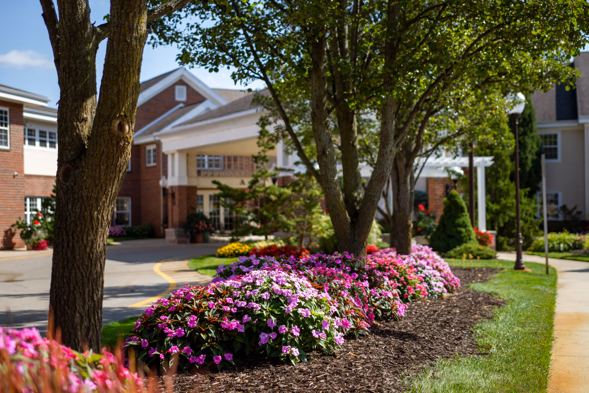 A landscaped outdoor area with colorful flower beds and trees in front of a brick and white senior living facility building under a clear blue sky.