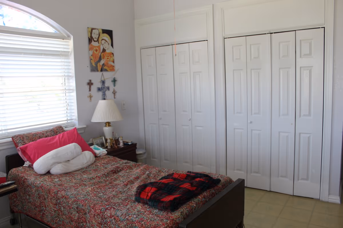 A bedroom with a single bed covered in a patterned bedspread and a red and black plaid blanket folded at the foot. There is a white pillow and a pink pillow on the bed. Next to the bed is a nightstand with a white lamp, framed photos, and small decorative items. On the wall above the nightstand are several crosses and a religious painting. A window with white blinds is on the left side, and two sets of white double-door closets are on the right wall.