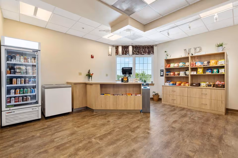 Interior view of a small convenience store area in a senior living facility with a refrigerated display case filled with beverages, a freezer, a wooden counter with snacks, and a wooden shelving unit stocked with various bags of chips. There is a window with a valance and some potted plants on the floor and shelves.