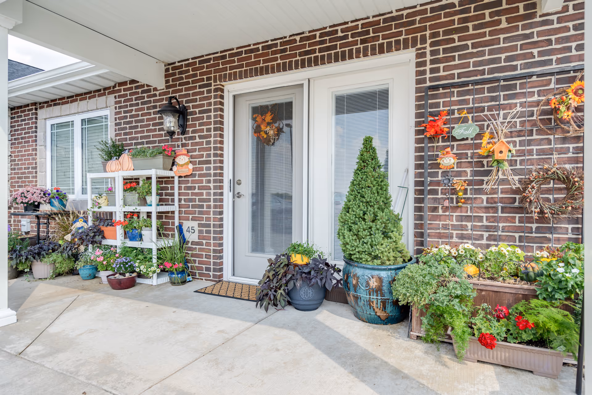 Brick-front entrance with double glass doors flanked by numerous potted plants, shelving of flowers and autumn decorations.