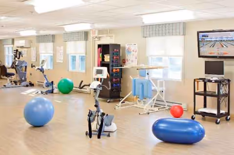 Bright indoor fitness room in a senior facility with exercise equipment and a wall-mounted TV.