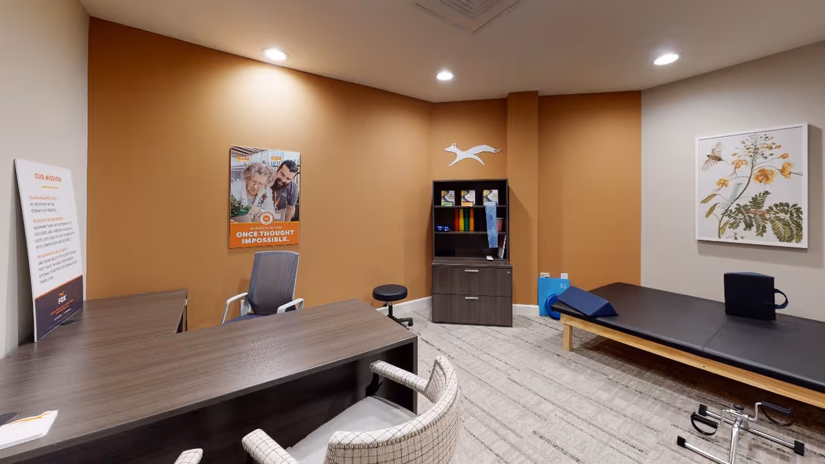A therapy or consultation room with a wooden desk and two chairs in the foreground, a gray office chair behind the desk, a black stool, a cabinet with therapy supplies, and a padded therapy table with a blue wedge cushion. The walls are painted in beige and orange tones, with a framed botanical print and a motivational poster on the walls.