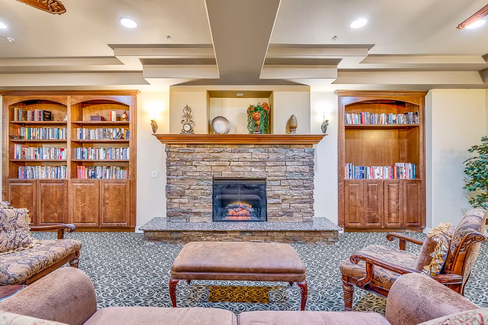 A cozy senior living room with a stone fireplace in the center, flanked by two wooden bookshelves filled with books. The room has patterned carpet, upholstered armchairs, and a cushioned ottoman in front of the fireplace. Decorative items are placed on the fireplace mantel and the walls have sconces providing warm lighting.
