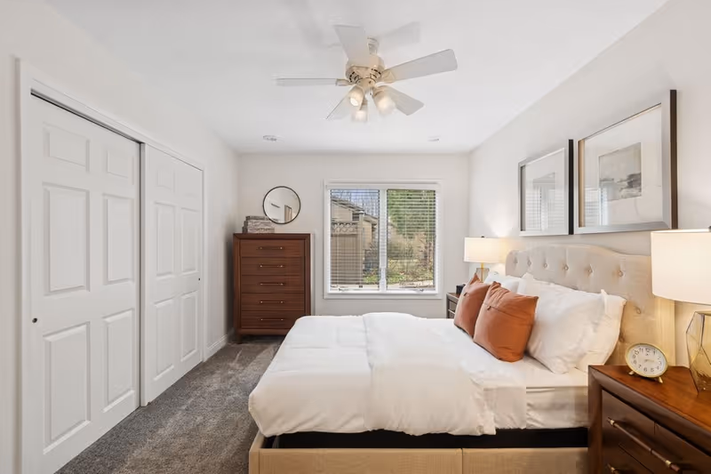 A bright and neatly arranged bedroom with a large bed featuring white bedding and three brown pillows. The room has a beige upholstered headboard, two wooden nightstands with lamps, a round clock, and framed artwork above the bed. There is a window with blinds letting in natural light, a tall wooden dresser with a round mirror above it, and a closet with white sliding doors. The ceiling has a white fan with lights.