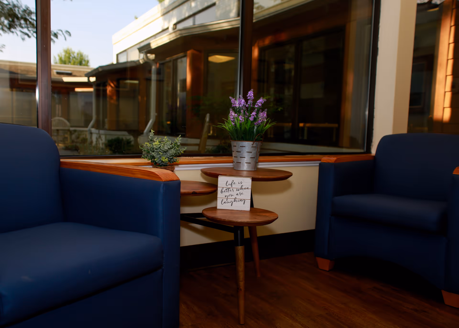 Two blue armchairs flanking a small wooden side table with potted plants and a decorative sign in front of a large window showing an outdoor courtyard.