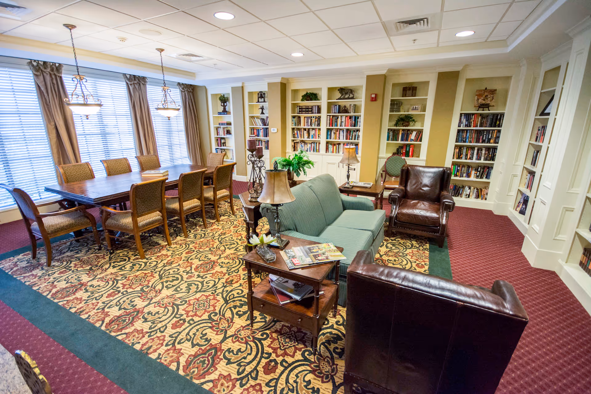 A cozy and well-lit common area featuring a large wooden table with eight chairs near windows with curtains, a green upholstered sofa, two brown leather armchairs, side tables with lamps, and built-in bookshelves filled with books and decorative items. The room has a patterned carpet and ceiling lights.