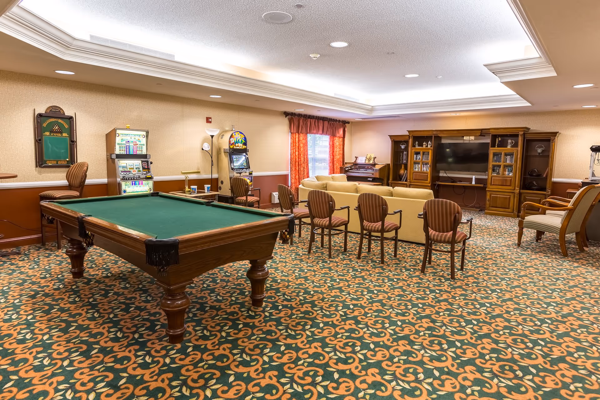 A spacious common room with a pool table in the foreground, a seating area of sofas and chairs facing a large TV and entertainment center, and game machines and a piano along the back wall.