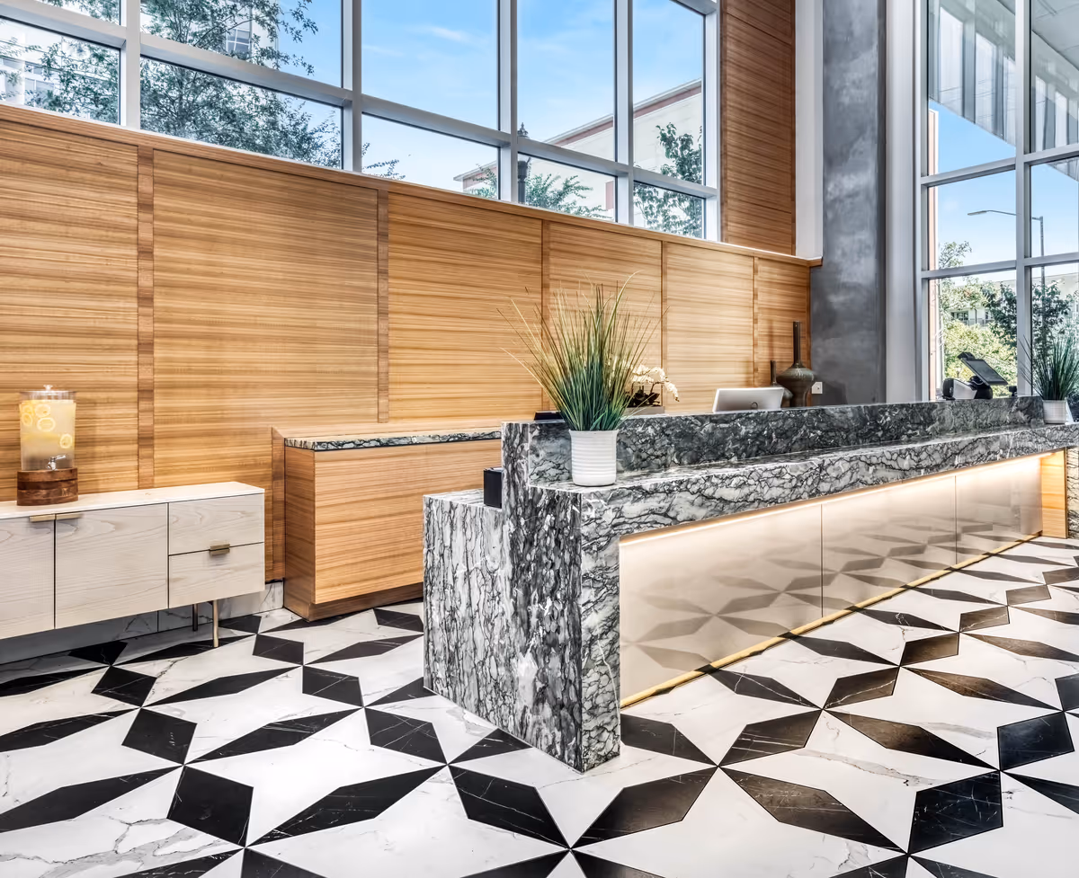 Modern reception area with a marble front desk, geometric black and white tiled floor, wooden panel walls, large windows letting in natural light, and decorative plants on the desk.