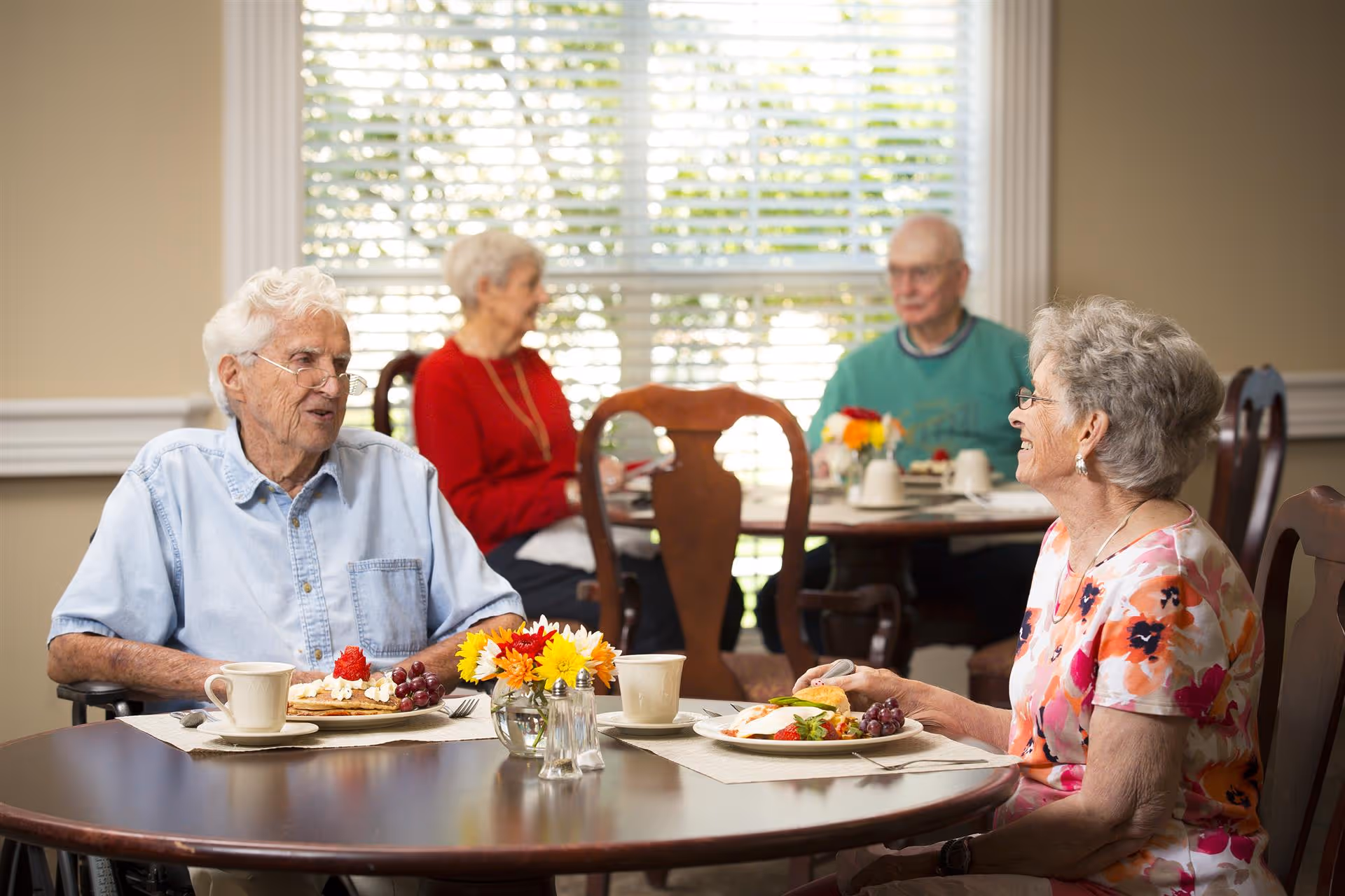 Elderly residents seated at round tables in a bright dining room, enjoying a meal and conversing.