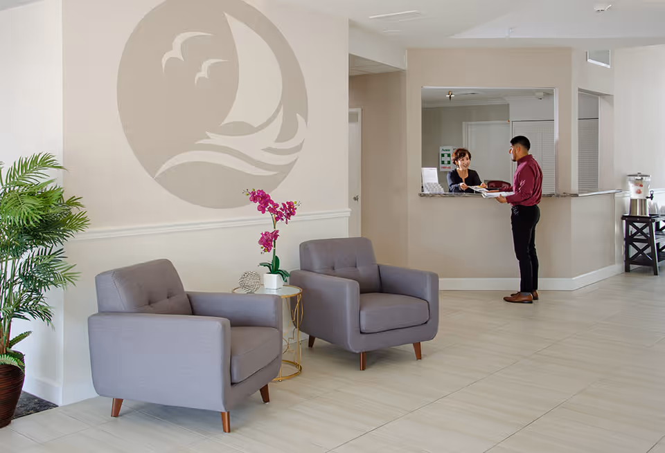 A reception area in a senior living facility with two gray armchairs and a small round table with a purple orchid plant. A man is standing at the reception desk talking to a woman behind the counter. There is a large wall decoration of a sailboat and birds on the wall behind the chairs, and a potted plant is placed to the left.