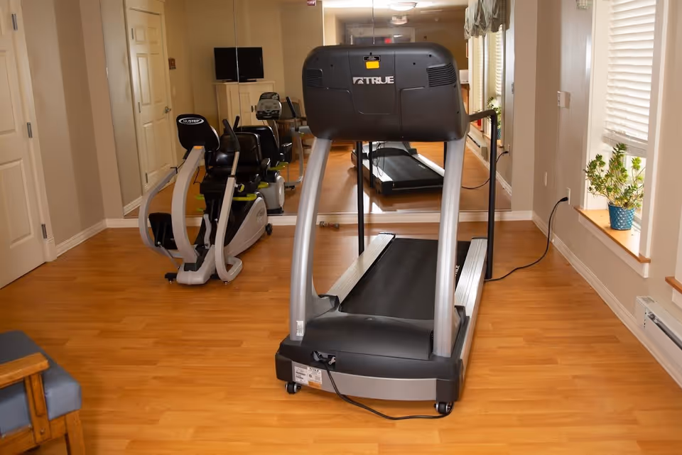 Exercise room with a treadmill and a recumbent exercise bike on a wooden floor. There is a large mirror on the wall reflecting the equipment and a window with blinds and a potted plant on the windowsill. A TV is mounted on a cabinet in the background.