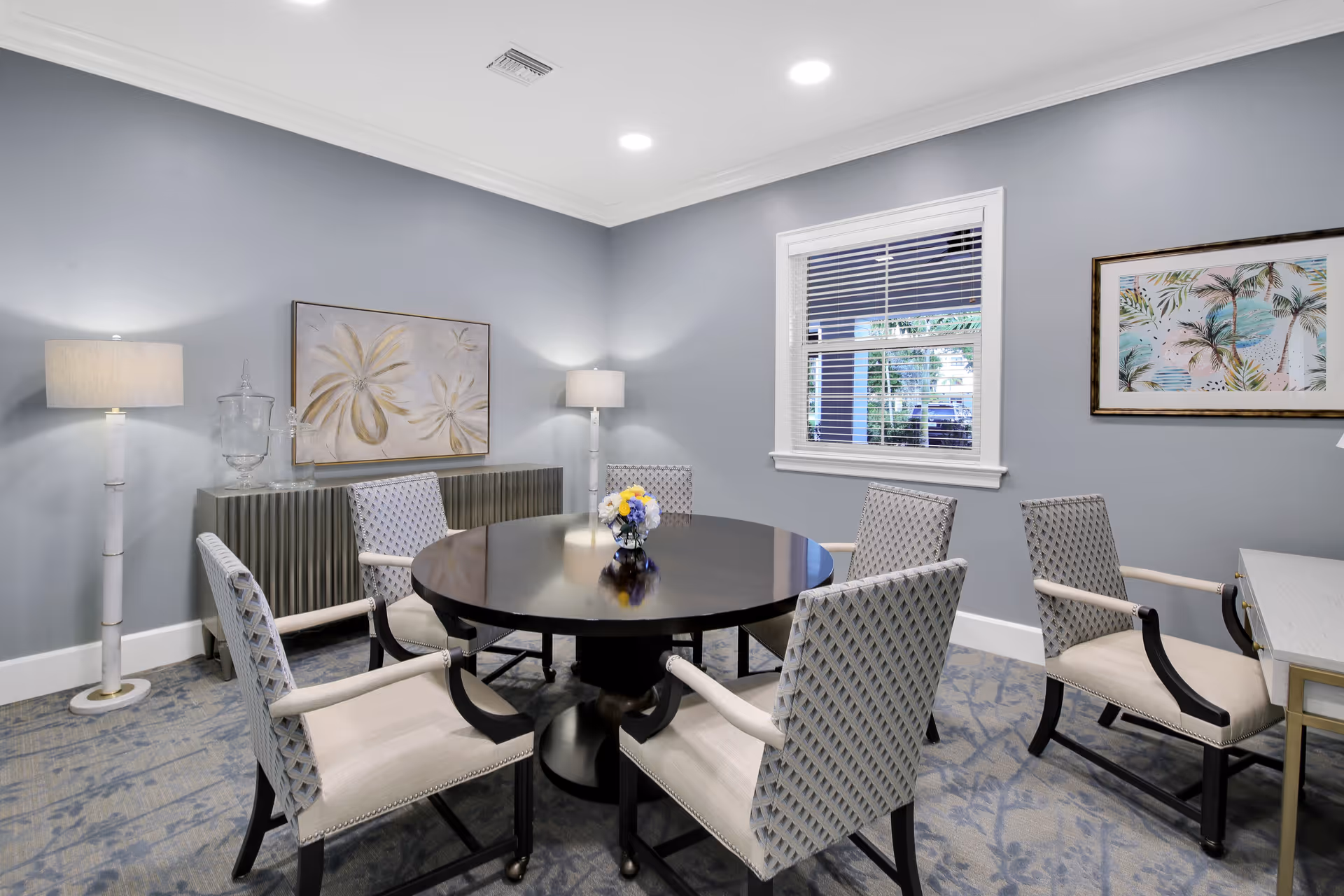 A well-lit room with a round dark wooden table surrounded by six cushioned chairs with patterned backs. The walls are painted light gray and decorated with two framed artworks, one featuring palm trees and the other floral designs. Two floor lamps with white shades stand on either side of a sideboard against the wall. A window with white blinds lets in natural light.