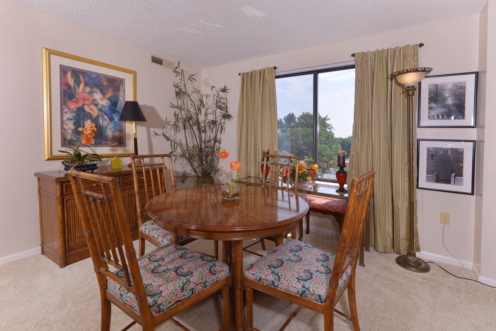 A dining room with a round wooden table surrounded by four wooden chairs with patterned cushions. Behind the table is a wooden sideboard with a lamp, a plant, and a framed colorful floral painting on the wall. To the right, there is a large window with green striped curtains, a bench with a red cushion, decorative candles, and framed black and white photos on the wall. A tall floor lamp stands in the corner.