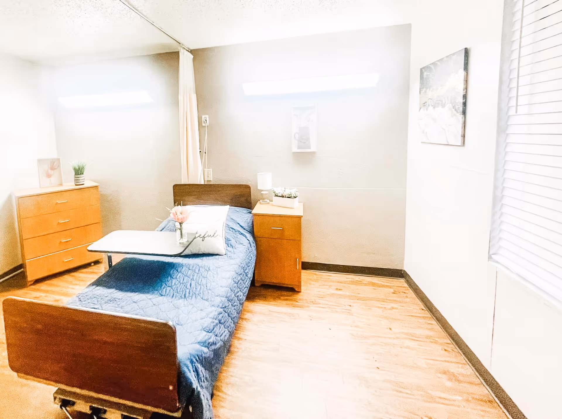 Well-lit nursing facility bedroom with a single bed, bedside table, dresser, and window blinds.