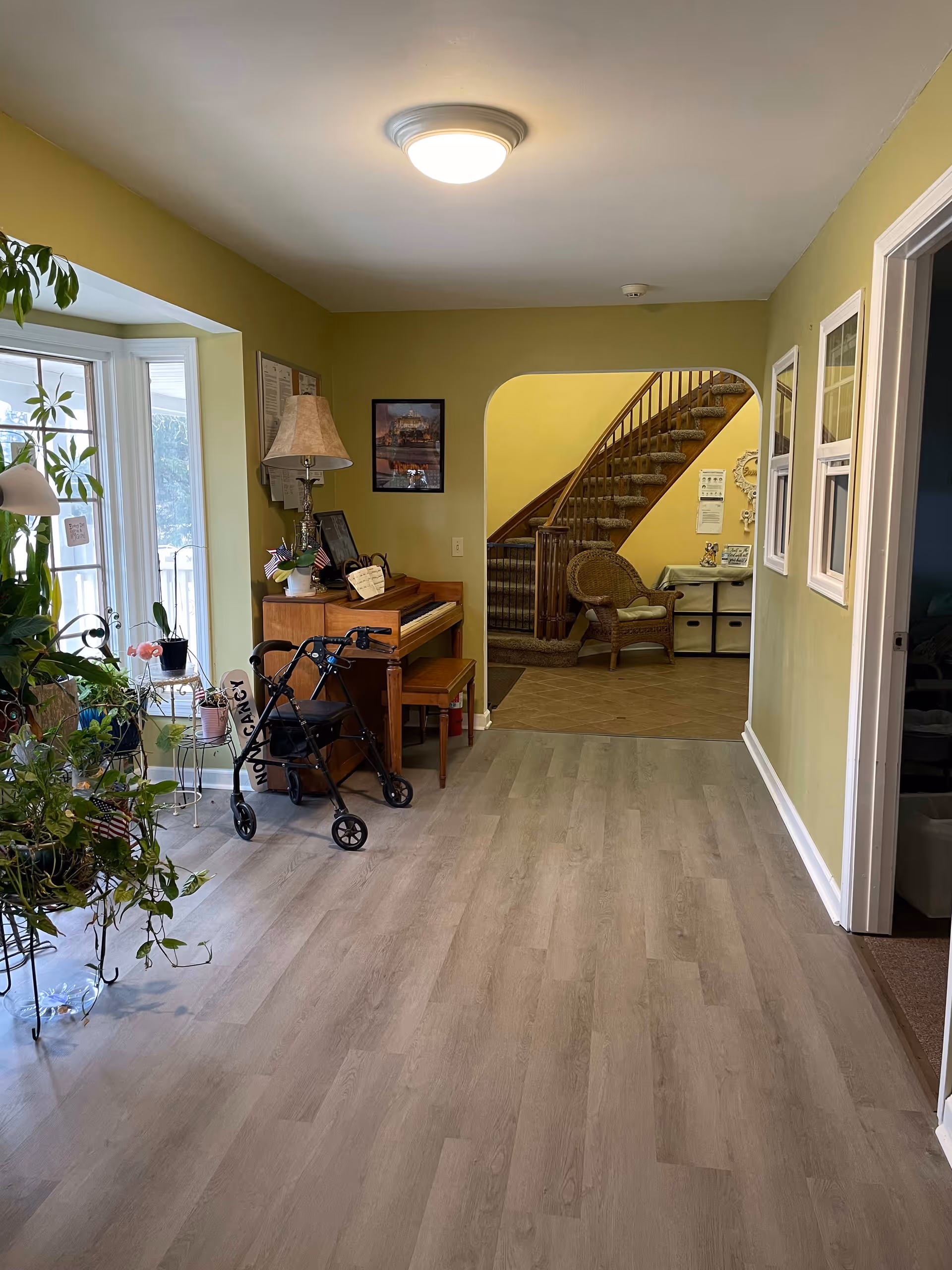 Interior view of a senior living facility hallway with light green walls and light wood flooring. On the left side, there is a bay window with several potted plants on stands. A wooden piano with a lamp and small American flags is against the wall. A black walker with a 'NOT A CHAIR' sign is positioned near the piano. In the background, an arched doorway leads to a carpeted staircase with a wicker chair and a small table with storage bins underneath. On the right side, there are two small windows and an open door leading to another room.