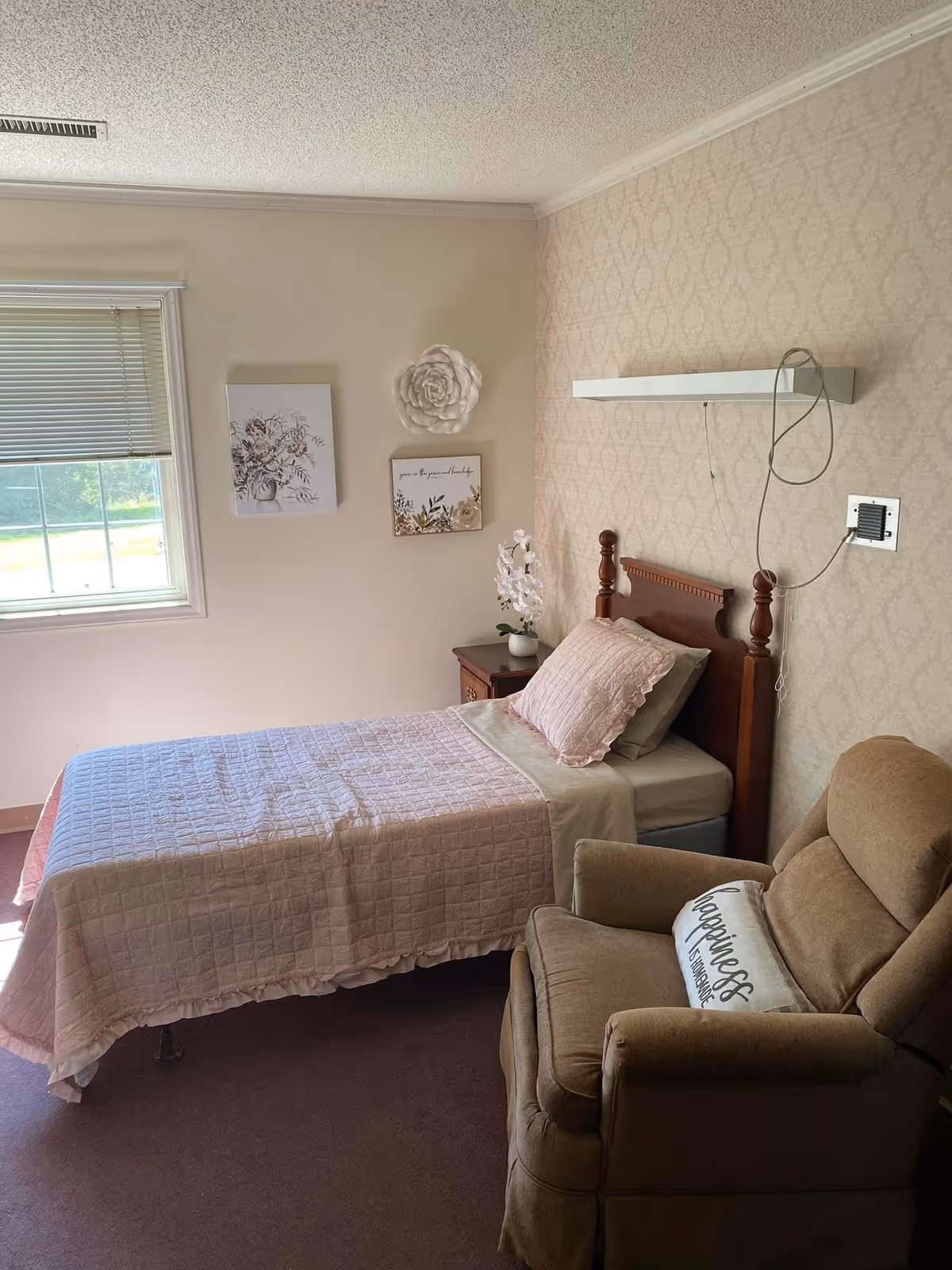 A cozy bedroom with a single bed covered in a pink quilt and matching pillow. Next to the bed is a wooden nightstand with a white orchid plant. On the wall above the nightstand are three decorative pieces: a floral painting, a white flower sculpture, and a framed quote. A beige recliner chair with a pillow that says 'Happiness is homemade' is positioned near the bed. A window with blinds lets in natural light, and the walls have a subtle patterned wallpaper.