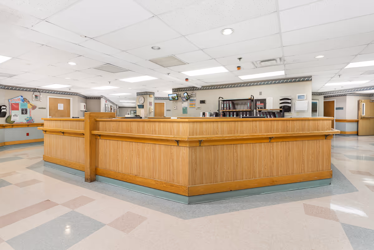 A spacious healthcare facility reception or nurse station area with a large wooden counter in the center. The room has a tiled floor with a geometric pattern, drop ceiling with fluorescent lighting, and walls decorated with bulletin boards, clocks, and mounted monitors. Several doors and hallways lead off from the area.