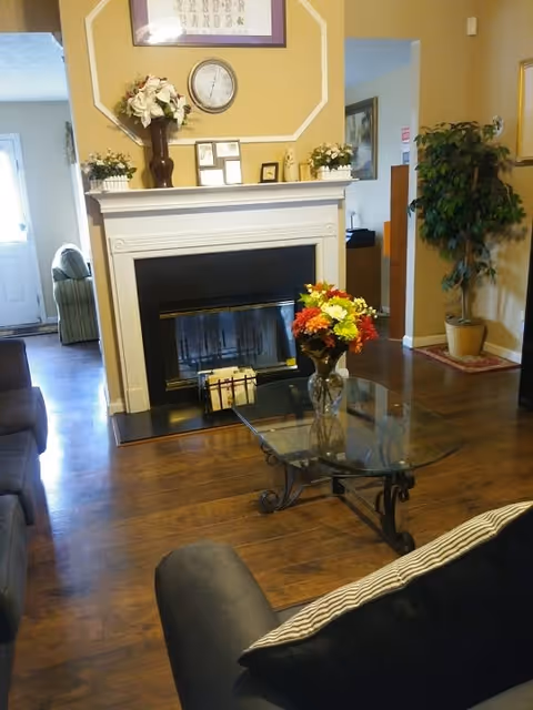 A cozy living room with a dark gray sofa and a glass coffee table with a vase of colorful flowers. There is a white fireplace with decorative items and a clock above it. The room has wooden flooring and beige walls, with a potted plant in the corner and an open doorway leading to another room.