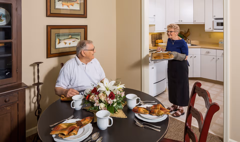 An elderly woman brings a tray of baked goods from a kitchen while an elderly man sits at a dining table set with plates and a floral centerpiece.
