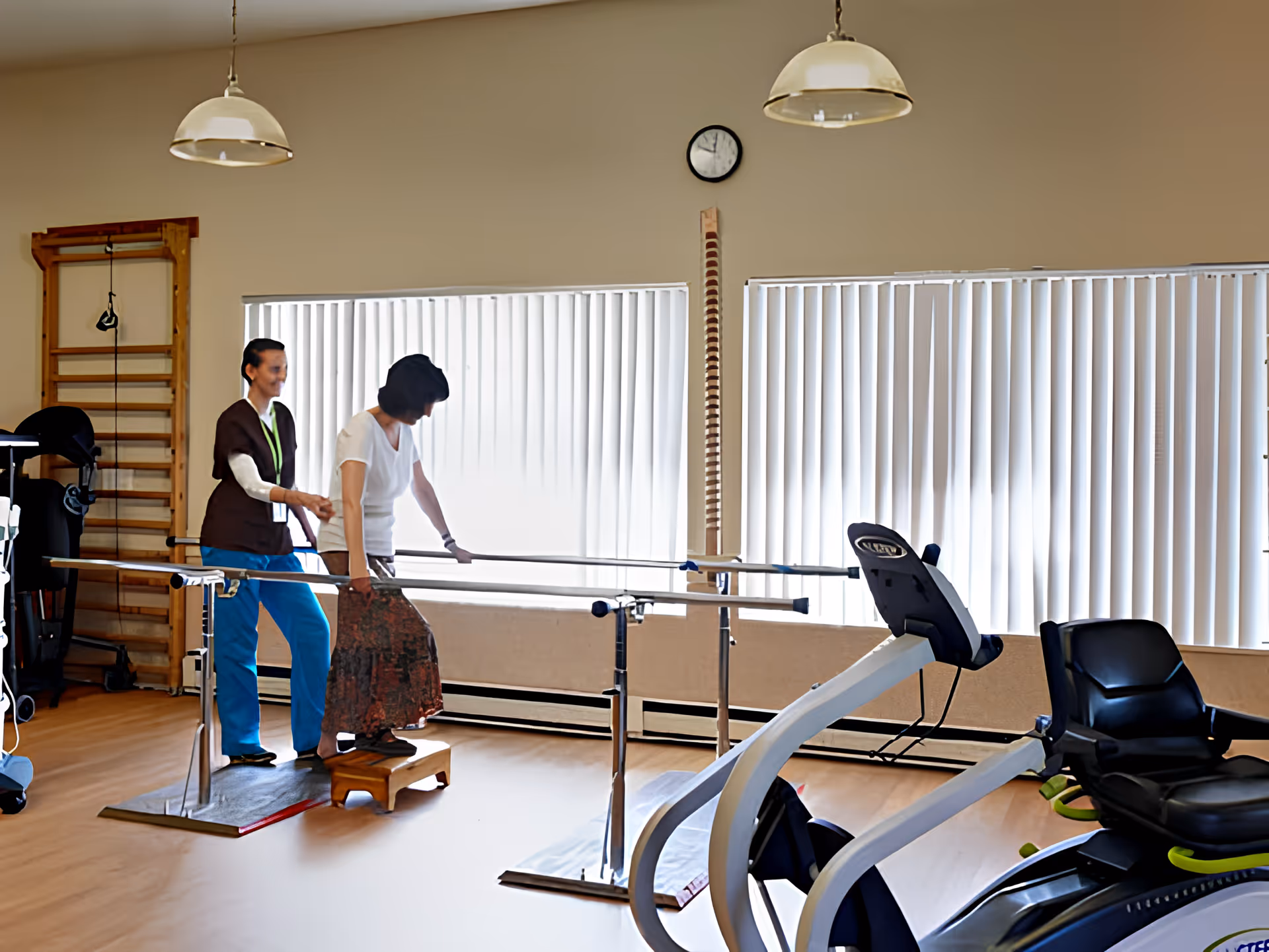 A rehabilitation gym room where a staff member assists a resident walking between parallel bars.