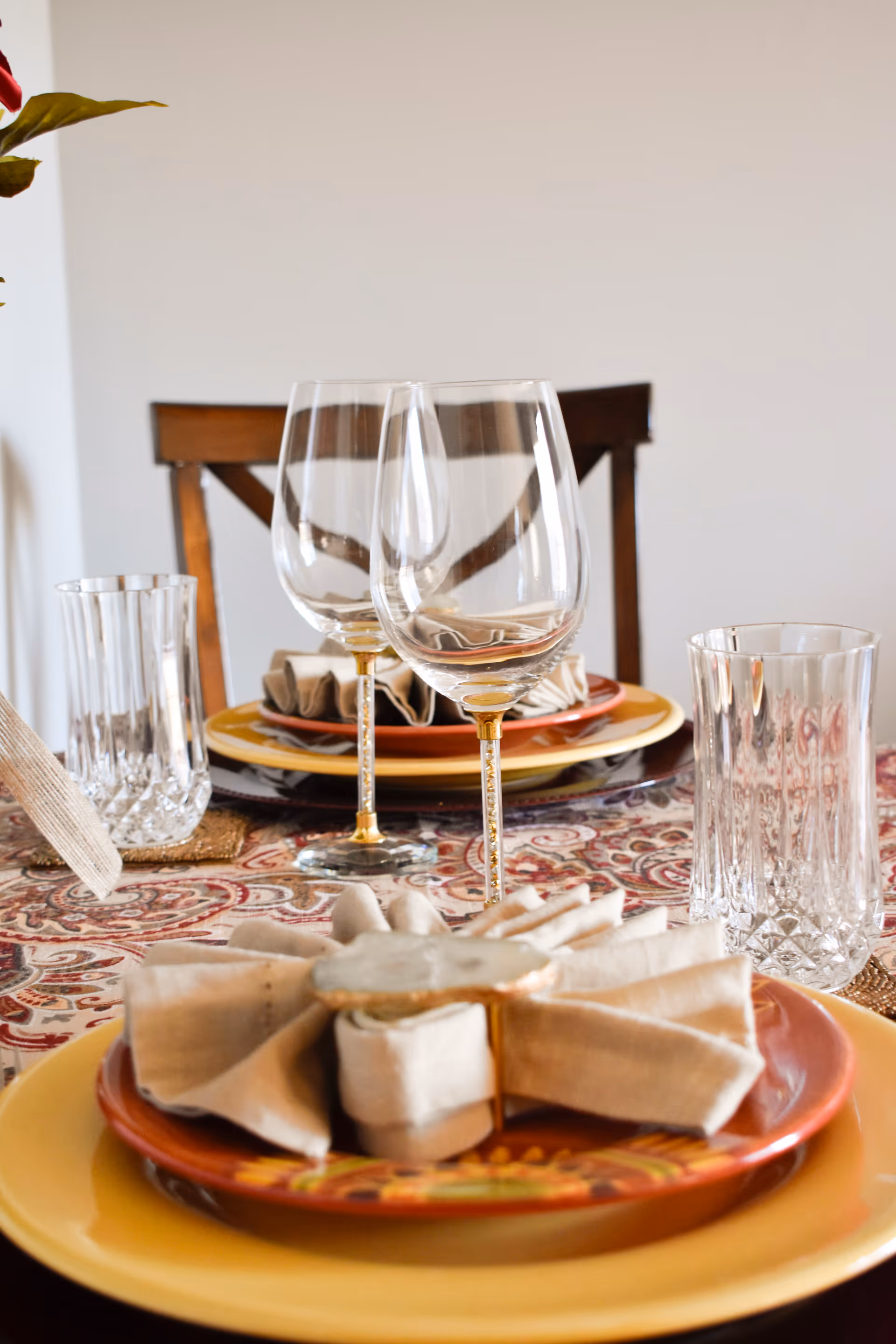 Close-up of a dining table set with yellow and orange plates, beige cloth napkins with napkin rings, two empty wine glasses, and two clear water glasses on a patterned tablecloth with a wooden chair in the background.