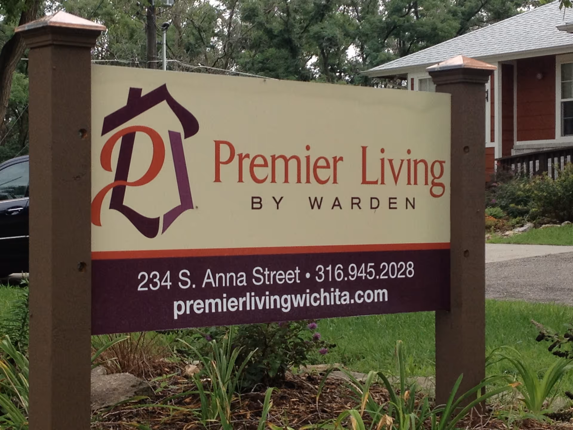 Outdoor sign for Premier Living by Warden, LLC, located at 234 S. Anna Street with phone number 316.945.2028 and website premierlivingwichita.com, surrounded by grass and plants with a house and trees in the background.