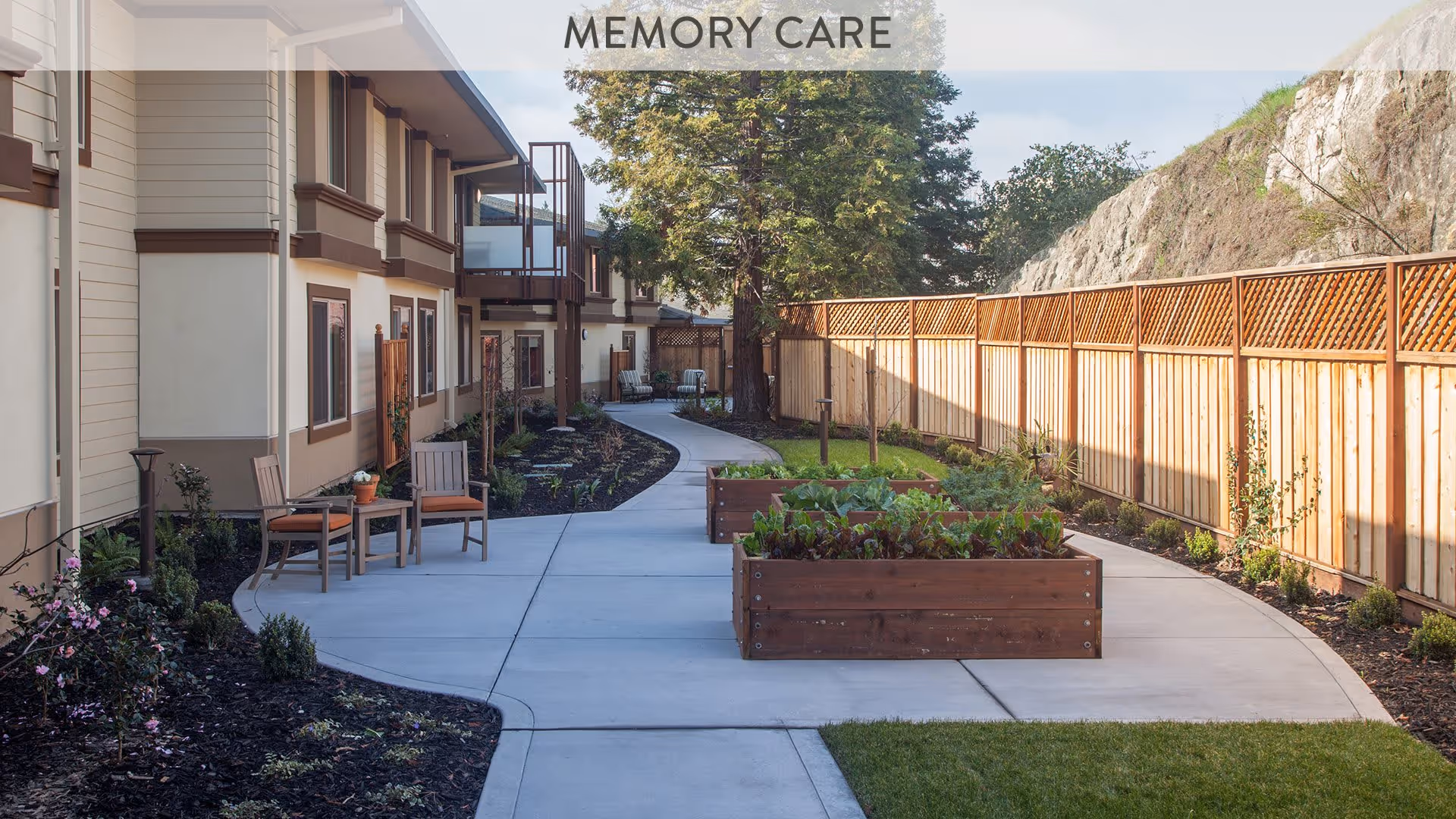 Outdoor garden area at Atria Walnut Creek with a paved walkway, wooden raised garden beds with plants, wooden fence on the right, and chairs along the building on the left. Trees and a rocky hill are visible in the background under a clear sky.