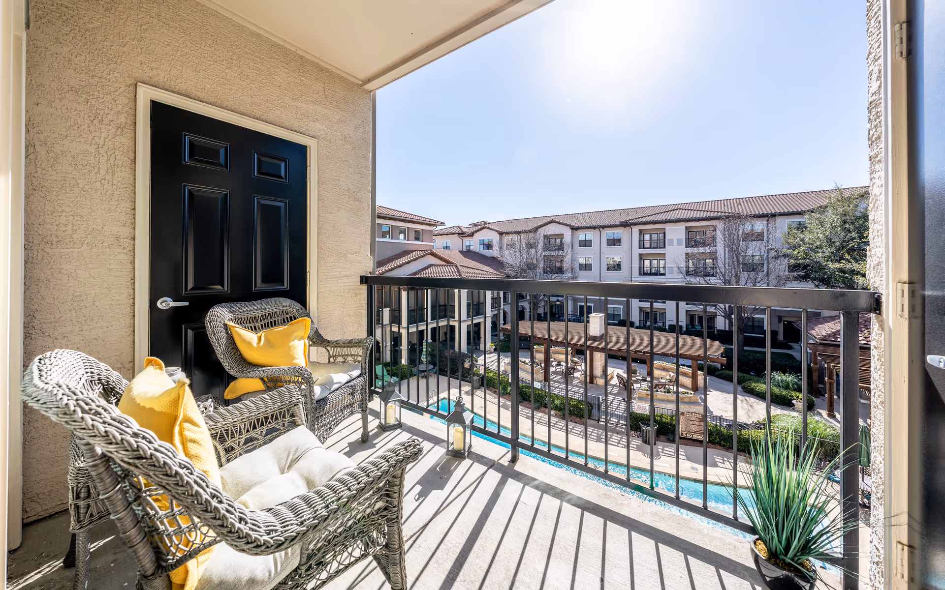 Balcony with two wicker chairs featuring yellow cushions, a black door, and a view overlooking a courtyard with a pool, seating area, and multi-story residential building under a clear sky.